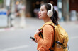 © Tj - Young Asian woman wearing headphones, holding smartphone, and carrying backpack while walking in urban street, enjoying travel and lifestyle