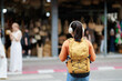 © Tj - Young female traveler with a yellow backpack and headphones exploring a vibrant street market, immersing in local culture while walking and listening to music