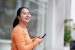 © Tj - Young smiling Asian woman confidently walking outdoors while holding a smartphone, communicating, connecting, and enjoying modern urban lifestyle and digital technology