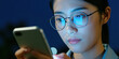 © Vadym - Young asian woman with glasses uses phone at night. Blue screen light reflects on her face. She texts or browses online. Woman looks serious, focused on mobile device.