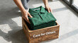 © aura - ​A person placing stacked green polo shirts into a wooden donation box with 'Care for Others' written on it.