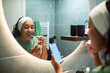 © Marko Geber - Woman brushing teeth while using smartphone in bathroom mirror