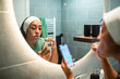 © Marko Geber - Woman brushing teeth and using smartphone in home bathroom mirror