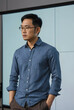 © Pixel-Shot - Thoughtful asian man in glasses and blue shirt with rolled sleeves standing in front of a blank projection screen, He is looking to the side with a serious expression, contemplation or deep thought