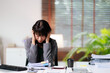 © Matawut - Asian businesswoman suffering from a stress-induced headache and burnout, grappling with overwhelm, overwork, and exhaustion at her office desk filled with documents and files