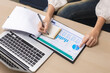 © Ratirat - Business person writing in notebook while reviewing chart and graph on desk next to laptop performing careful data analysis for future corporate strategy and financial planning