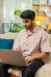 © Andrii Iemelianenko - Indian man on home sofa works on laptop with focused face typing checking project result for remote job indoors. Arabian guy on home sofa reviews online task with calm smile in living room workspace.
