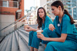 © Zamrznuti tonovi - Tired female doctors in medical scrubs sitting on outdoor stairs, drinking coffee, and discussing work during a break