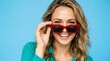 © samia - Playful young woman with wavy blonde hair wearing teal blouse holding bold red sunglasses above her face, smiling brightly with perfect teeth against solid blue background, summer fashion accessory,