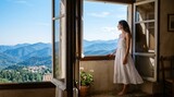 A serene woman in a flowing white summer dress stands barefoot at a tall open wooden window, gazing out at distant blue mountain ranges and a lush green valley village under a clear sunny blue sky.