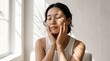 © m0lydar - Young Asian woman with glowing natural skin gently touching her face while enjoying soft morning sunlight in a peaceful indoor setting