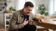 © Taras Roshchuk - A man sits at a wooden table in a bright living room. He is holding his chest with one hand and appears to be in discomfort. The room has plants and natural light