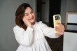 © timoha.ig - Young woman smiling while holding smartphone for a video call in a modern home setting. Social media influencer marketing and personal branding