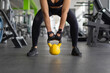 © Anastasiya - Closeup of young Black woman gripping yellow kettlebell between her legs, demonstrating functional strength, proper stance and preparation for a gym workout