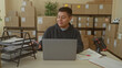 © Krakenimages.com - Man at laptop smiling and typing while glancing at phone amid stacked parcels in a building; small business contentment.