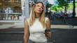 © Krakenimages.com - Young woman with long blond hair holding a takeaway coffee cup and wearing clear glasses on a city street in front of a cafe storefront; annoyance waiting.