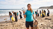 © Alexander - Young woman holding collected plastic bottles with volunteer team cleaning beach in background, recycling and sustainability awareness