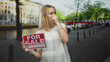 © Krakenimages.com - Blonde woman holding for sale sign pinches nose with hand and grimaces while standing on a cobblestone street by a canal; sale frustration disgust.
