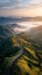 © Andi - Aerial view of a winding road through lush green rolling hills covered in morning mist at sunrise