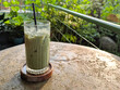 © Maule - Iced matcha latte with a black straw in a ribbed glass on a wooden coaster, sitting on a textured stone table with a lush green garden and warm sunlight in the background.