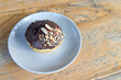 © Maule - A single chocolate-covered donut topped with chopped almonds and slivers, plus chocolate chips, served on a grey plate on a rustic wood table.