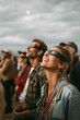 © Lady O - Group of people gathered outdoors wearing eclipse glasses, looking up in awe, subtle smiles and wonder on their faces.