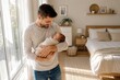© IrenKa - Smiling young father gently holding his sleeping newborn baby in a bright modern bedroom with natural window light, celebrating love, care and early parenthood