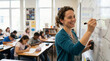 © Matos - Smiling teacher writing on whiteboard in classroom with students learning
