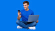 © Vyatcheslav - Smiling young man sitting cross legged with laptop and smartphone. Male multitasking on blue background