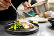 © pavel siamionov - A chef in a commercial kitchen plates a dish, pouring a creamy white sauce from a small metal pot over a grilled salmon fillet served with green beans on a sleek black plate