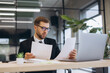 © anatoliycherkas - Young businessman reviewing financial reports in office, analyzing documents and working on laptop