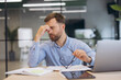 © anatoliycherkas - Man experiencing work burnout and fatigue, holding glasses while sitting at a desk with laptop and documents