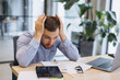 © anatoliycherkas - Young businessman experiencing work burnout and stress, holding head in hands at office desk with documents and laptop