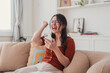 © Daniel - Young woman sitting on a sofa, looking at her smartphone and cheering with a fist pump, expressing excitement and joy after receiving good news or achieving success