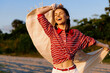 © SHOTPRIME STUDIO - A young woman in a red striped shirt twirls on a sunlit beach, scarf fluttering behind her, hair loose, face lit with a bright, carefree smile, capturing a joyful lifestyle moment.