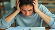 © realone952 - A woman sits at a table with a headache and holds her head in her hands, over documents with calculations.