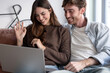© luciano - Happy couple relaxing on sofa at home, enjoying a video call on laptop together, waving goodbye to friends or family, maintaining connection and communication