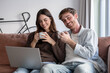 © luciano - Happy young couple enjoying a relaxing morning on the sofa at home, drinking coffee and tea while browsing internet content on a laptop computer