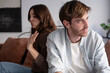© luciano - Young couple sitting back to back on a sofa at home after a fight, the woman gesturing while the man looks away, feeling frustrated and distant