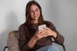 © luciano - Young adult woman smiling and enjoying her phone, connecting with others, browsing online, and relaxing comfortably while sitting on a chair indoors