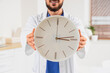 © Pixel-Shot - Doctor in white coat holding a large wall clock medical appointment scheduling and time management in healthcare