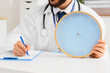 © Pixel-Shot - Doctor in white coat holding a large blue clock and writing on a clipboard, medical appointment time management, the physician is simultaneously writing on a clipboard with a blue pen