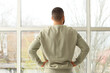 © Pixel-Shot - Young man looking out large window at home, back view