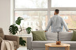 © Pixel-Shot - Young man behind sofa looking out large window at home, back view