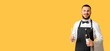 © Pixel-Shot - Smiling young male waiter in black apron holding a wine bottle and empty glass on a yellow background, He wears a black bow tie and has a checkered towel draped over his arm, standing