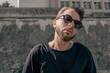 © Valeria Vaskovats - Stylish young man in black contemporary clothing and round sunglasses posing outdoors against a modern concrete urban background in natural daylight.