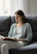 © Pixel-Shot - Young woman reading a book on a comfortable gray sofa at home, enjoying quiet leisure time and relaxation