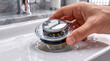 © Marina - A hand places a new polished chrome pop-up drain stopper plug unit into a ceramic bathroom washbasin sink opening.