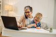 © standret - Elderly woman with short gray hair multitasking on a laptop while talking on the phone, young boy coloring beside her at a modern home office desk