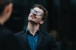 © qunica.com - Young businessman in a blazer and glasses looks upward with a thoughtful expression. A close urban portrait of a professional man gazing skyward in a suit and shirt.